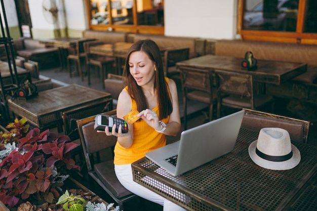 Mujer en la cafetería de la calle al aire libre sentada con una computadora portátil, tiene una terminal de pago bancaria moderna inalámbrica para procesar la adquisición de pagos con tarjeta de crédito