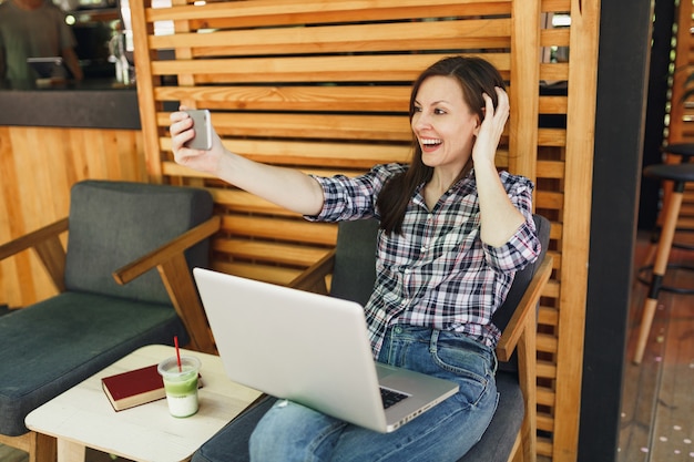 Mujer en la cafetería de verano de la calle al aire libre de madera sentado con la computadora portátil, haciendo selfie shot en el teléfono móvil