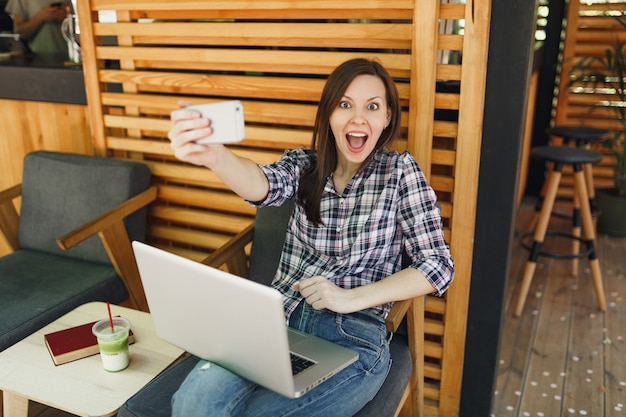 Mujer en la cafetería de verano de la calle al aire libre de madera sentado con la computadora portátil, haciendo selfie en teléfono móvil, relajándose durante el tiempo libre. Oficina móvil