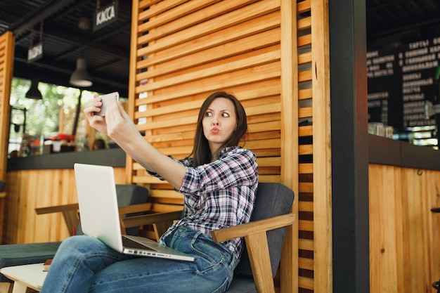 Mujer en la cafetería de verano de la calle al aire libre de madera sentado con la computadora portátil, haciendo selfie en teléfono móvil, relajándose durante el tiempo libre. Oficina móvil