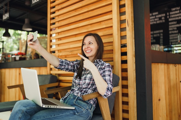 Mujer en la cafetería de verano de la calle al aire libre de madera sentado con la computadora portátil, haciendo selfie en teléfono móvil, relajándose durante el tiempo libre. Oficina móvil