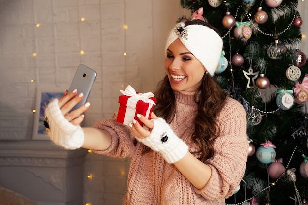 Mujer con caja de regalo comunicándose por teléfono móvil smartphone llamada en línea. Celebración remota de Feliz Navidad