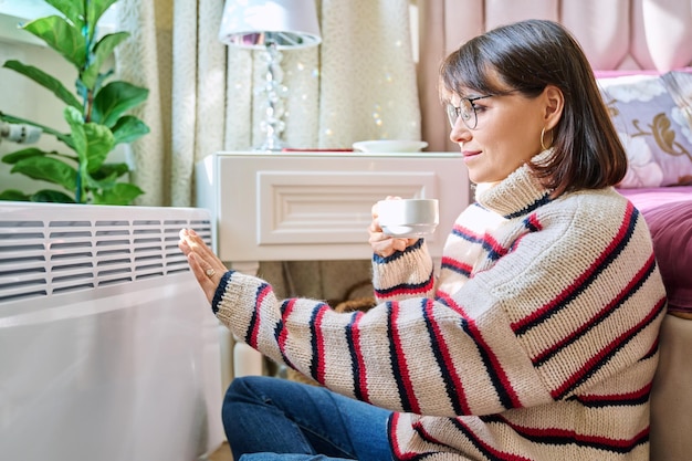 Foto mujer calentándose en el dormitorio cerca del calentador eléctrico