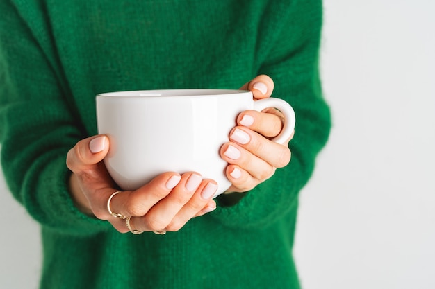 Foto mujer en cálido suéter verde de lana está sosteniendo la taza blanca en sus manos con té. simulacros de diseño de invierno. estilo minimalista.