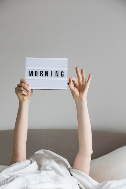 Mujer en la cama debajo de las sábanas sosteniendo un cartel matutino