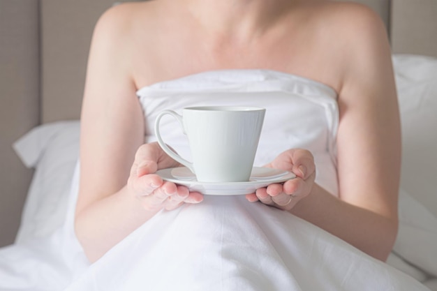 Mujer en la cama sostiene una taza blanca sobre un platillo en sus manos