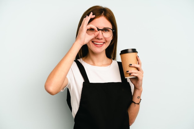 Mujer camarera de restaurante sosteniendo un café para llevar aislado en fondo azul emocionada manteniendo el gesto correcto en el ojo.