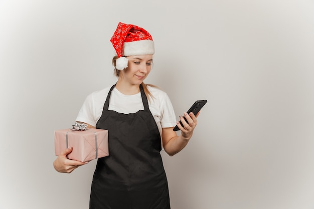 Mujer camarero con gorro de Papá Noel con un teléfono y un regalo en sus manos sobre un fondo gris aislado