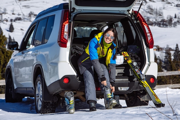 Mujer cambiarse de botas para esquiar sentado en el maletero del coche. día soleado. viaje por carretera de invierno