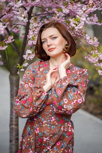 Una mujer camina por una calle de primavera. La niña disfruta del aroma de un árbol en flor. Hermosa mujer en un vestido con flor de cerezo sakura