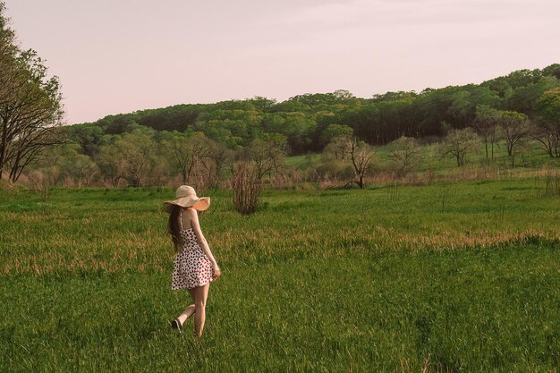 Una mujer camina por un campo con un vestido rosa y blanco y un sombrero.