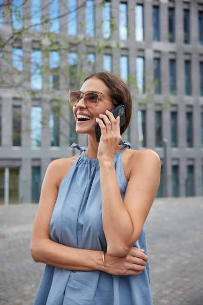 mujer camina en la ciudad urbanidad tiene conversación telefónica en itinerancia disfruta de charlas amistosas paseos durante el tiempo libre vacaciones de recreación usa un traje elegante gafas de sol explora un nuevo lugar