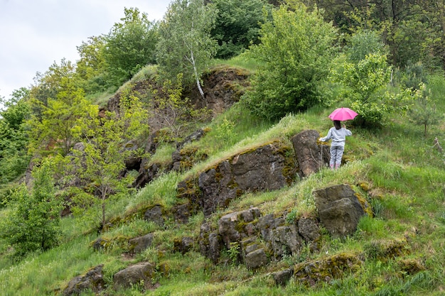 Una mujer camina bajo un paraguas en las montañas, entre las rocas cubiertas de vegetación.