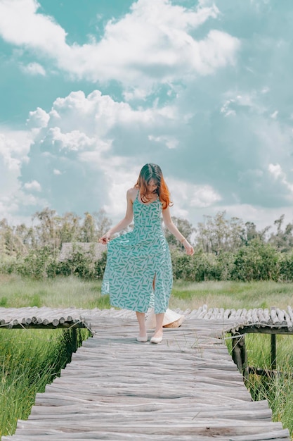 Una mujer camina sobre un puente de madera en un campo verde con un cielo nublado al fondo.