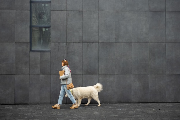 Una mujer camina con su perro blanco sobre el fondo de una pared gris al aire libre