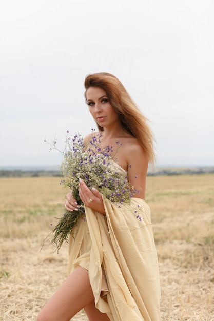 Mujer caminando en campo de hierba seca dorada. Belleza de retrato natural. Hermosa chica en un campo de trigo. Mujer joven con un vestido beige con un ramo de flores silvestres.