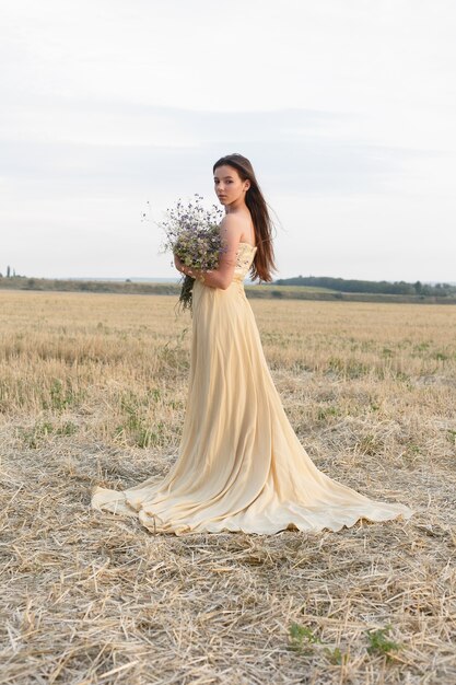 Mujer caminando en campo de hierba seca dorada. Belleza de retrato natural. Hermosa chica en un campo de trigo. Mujer joven con un vestido beige con un ramo de flores silvestres.