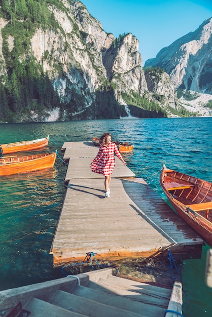 Mujer caminando por el muelle del lago de montaña con botes de madera en vestido rojo