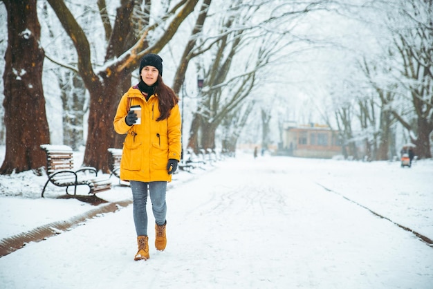 Mujer caminando por el parque de la ciudad nevada con cara pensante con taza de café calentar día frío