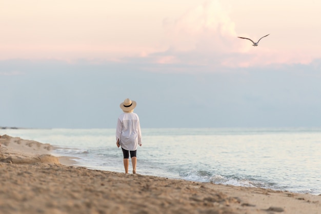 Mujer caminando por la playa al atardecer con teléfono celular y con sombrero de paja y soñando