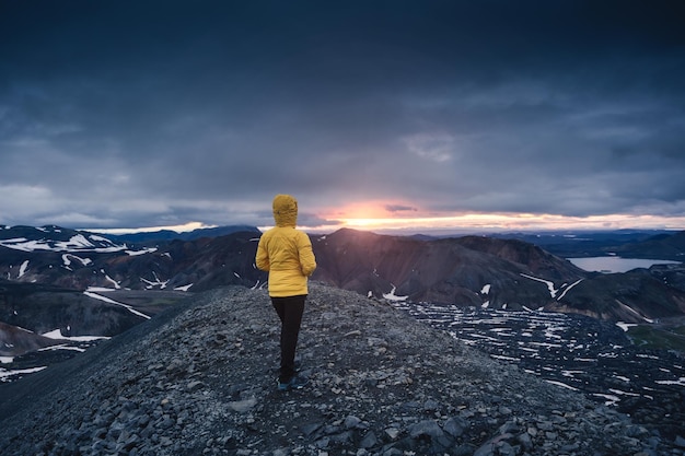 Mujer caminante con chaqueta amarilla parada en una montaña volcánica y sol de medianoche desde el sendero Blahnjukur en las tierras altas de Islandia