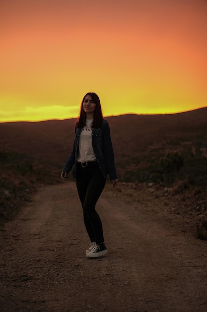 Una mujer se para en un camino de tierra frente a una puesta de sol.