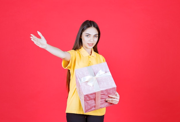 mujer con camisa amarilla sosteniendo una caja de regalo rosa e invitando a alguien a acercarse y tomarla.