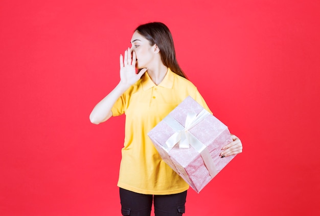 mujer con camisa amarilla sosteniendo una caja de regalo rosa y llamando a alguien.