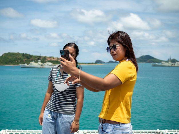 Una mujer con una camisa amarilla sostiene un teléfono para tomarse una selfie con una chica con una camisa gris con un fondo de agua azul y pequeñas montañas Es turismo después del brote del virus de la corona