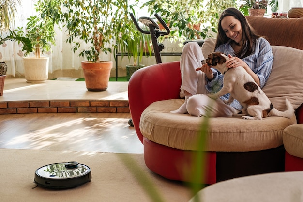 Mujer con camisa azul juega con perro, raza Jack Russell Terrier en casa en el sofá, aspiradora robótica en la alfombra