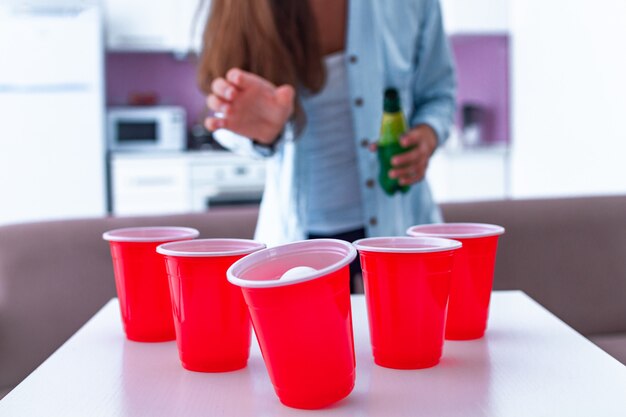 Mujer en camisa con bebidas divirtiéndose y disfrutando el juego de pong de cerveza en la mesa en casa