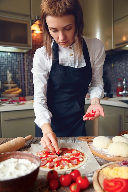 Mujer en camisa blanca y delantal poniendo los ingredientes en la pizza