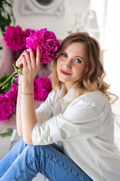 Mujer con camisa blanca y jeans azul, tiene ramo de peonías rosas en sus manos