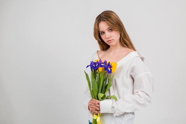 Una mujer con camisa blanca y jeans azules se sienta y sostiene un ramo de tulipanes e iris