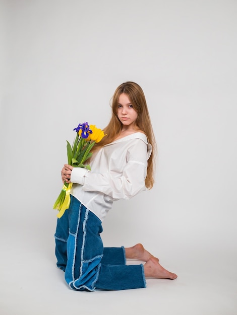 Una mujer con camisa blanca y jeans azules se sienta y sostiene un ramo de tulipanes e iris