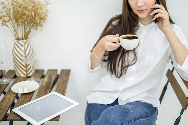 Una mujer con una camisa blanca y jeans se sienta en un café y bebe café Oficinista en la hora del almuerzo hablando por teléfono Gerente en una reunión de negocios en un restaurante