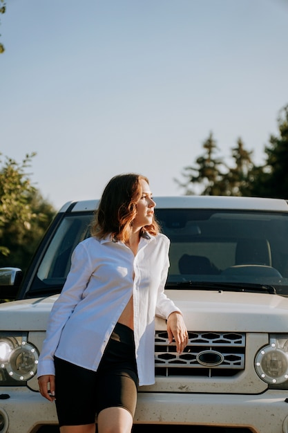 Una mujer con una camisa blanca junto a un automóvil blanco en la carretera. Un viaje a la naturaleza, descanso fuera de la ciudad. Foto vertical