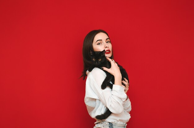 Mujer con camisa blanca y maquillaje brillante sobre fondo rojo, posando a la cámara con gato en manos