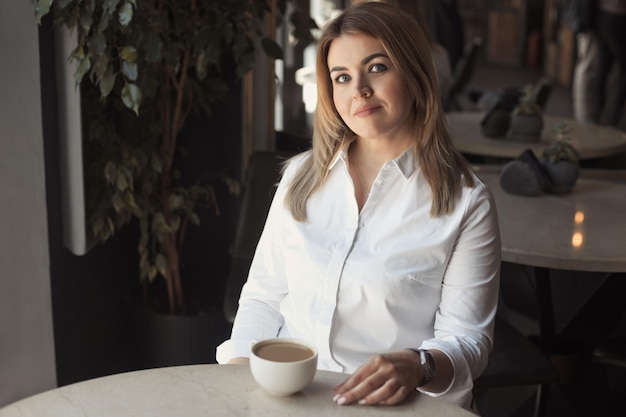 Mujer con camisa blanca de oficina con cabello rubio de 40 años bebiendo una taza de café en un café