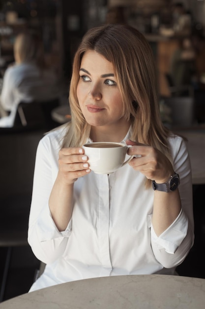 Mujer con camisa blanca de oficina con cabello rubio de 40 años bebiendo una taza de café en un café