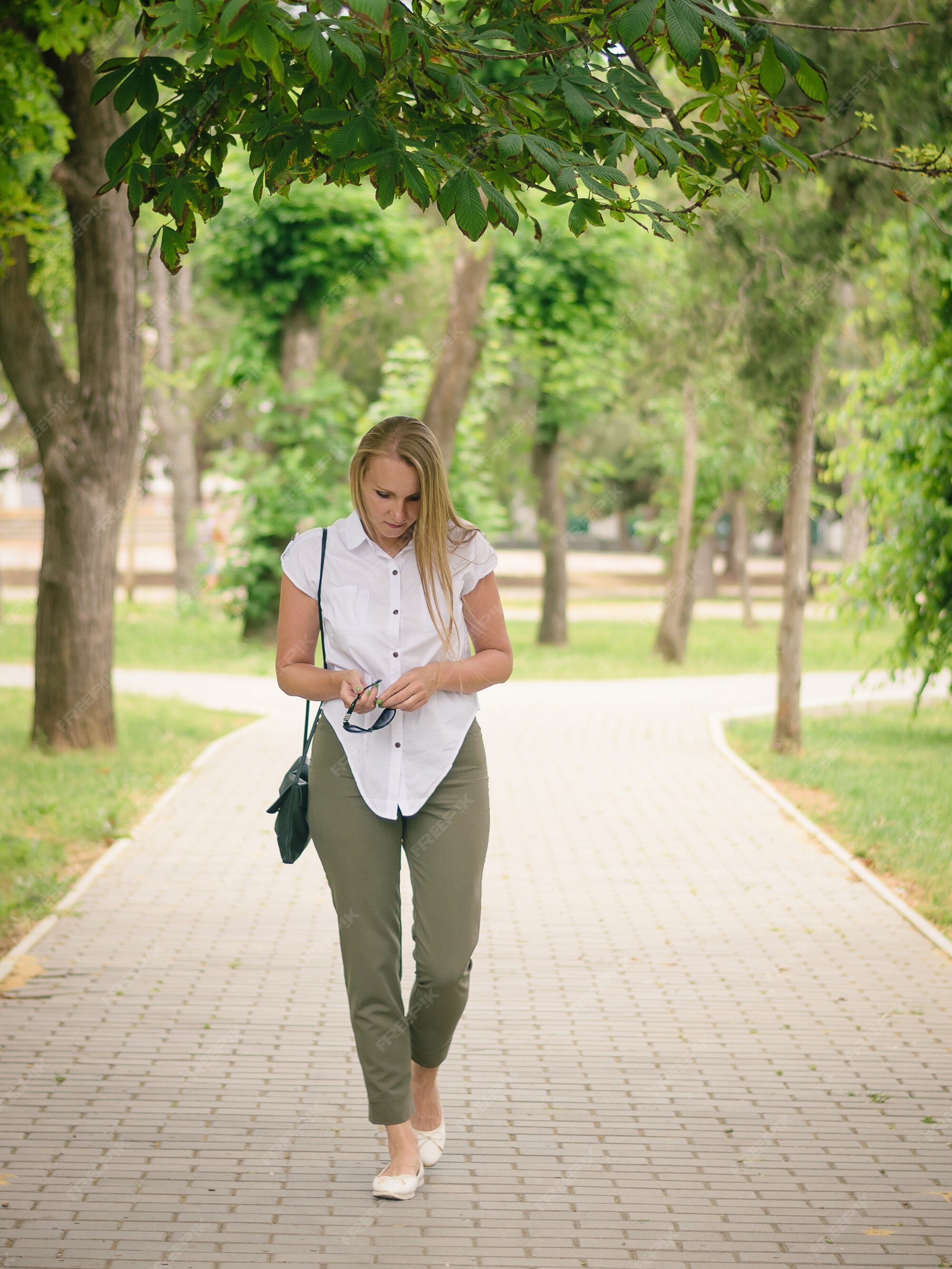 Mujer en camisa blanca y pantalón verde con gafas de sol en la mano caminando por el parque. | Foto Premium