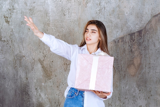 mujer con camisa blanca sosteniendo una caja de regalo rosa envuelta con cinta blanca, notando a su pareja y pidiéndole que venga a recibirla.