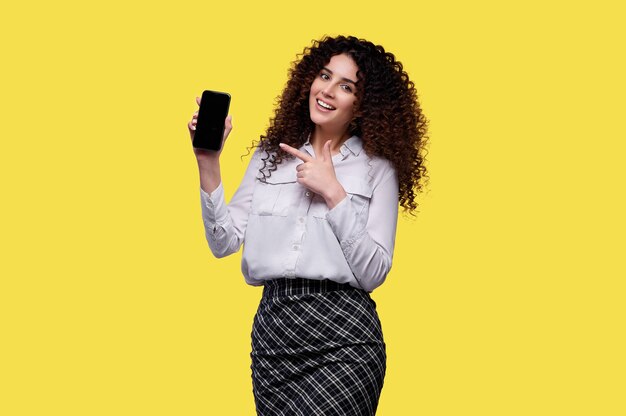 Mujer con camisa blanca tiene smartphone y apunta con el dedo índice a la pantalla en blanco. Empresaria sobre fondo amarillo aislado. Concepto de casino online