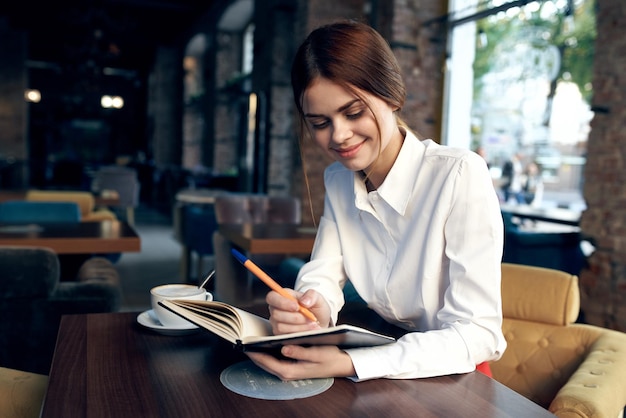 Una mujer en una camisa con un cuaderno y un bolígrafo en la mano se sienta en una mesa en un restaurante en una silla tapizada