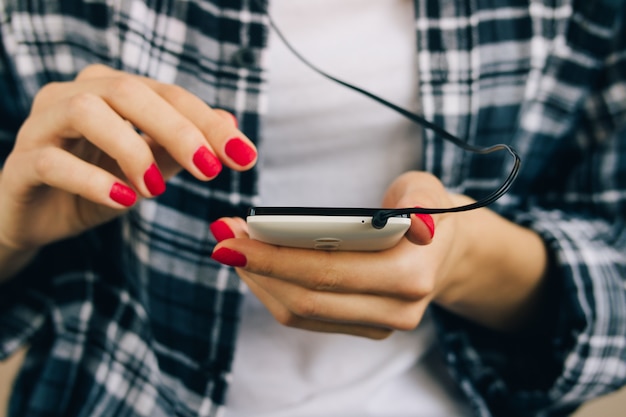 Mujer en camisa a cuadros y manicura roja con teléfono móvil blanco con auriculares conectados