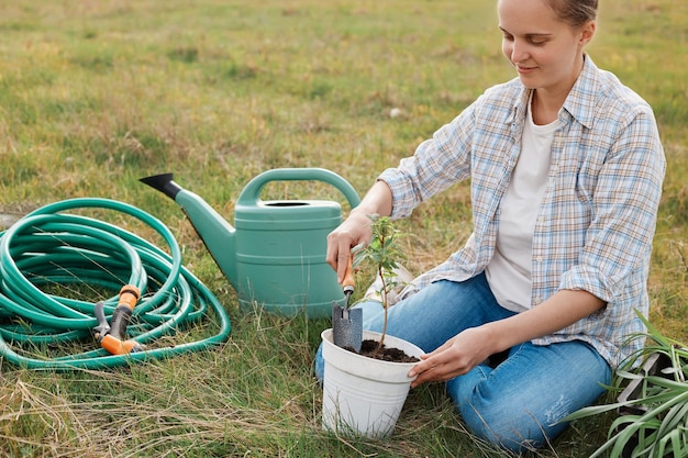 Mujer con camisa y jeans trabajando en el campo cavando plántulas de frambuesa de la olla sosteniendo una pala plantando un pequeño arbusto de bayas en el suelo rodeado de una lata de agua y una manguera de riego