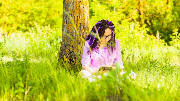 Una mujer con una camisa morada y anteojos lee una literatura debajo de un árbol, una niña descansa con un libro