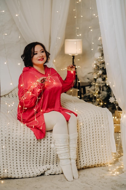 Una mujer con una camisa roja en una habitación decorada para Navidad está sentada en una cama Ambiente de Año Nuevo en casa