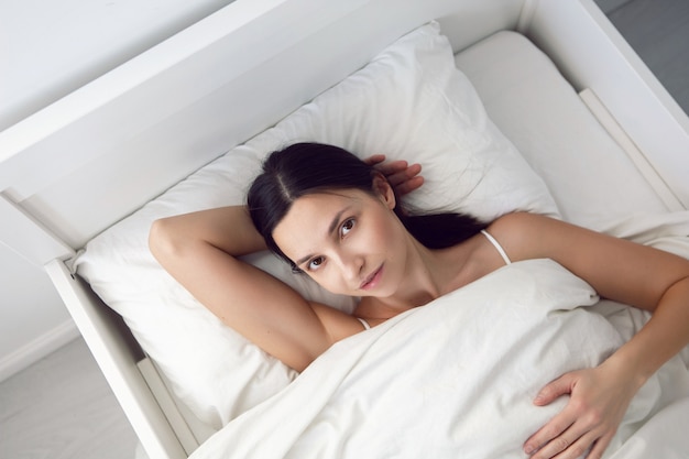 Mujer con una camiseta blanca está acostada en la cama y una almohada debajo de la manta en la habitación