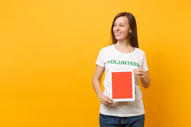 Mujer en camiseta blanca inscripción escrita título verde voluntario mantenga tablet pc computadora, pantalla vacía en blanco aislada sobre fondo amarillo. Ayuda de asistencia gratuita voluntaria, concepto de trabajo de gracia de caridad.